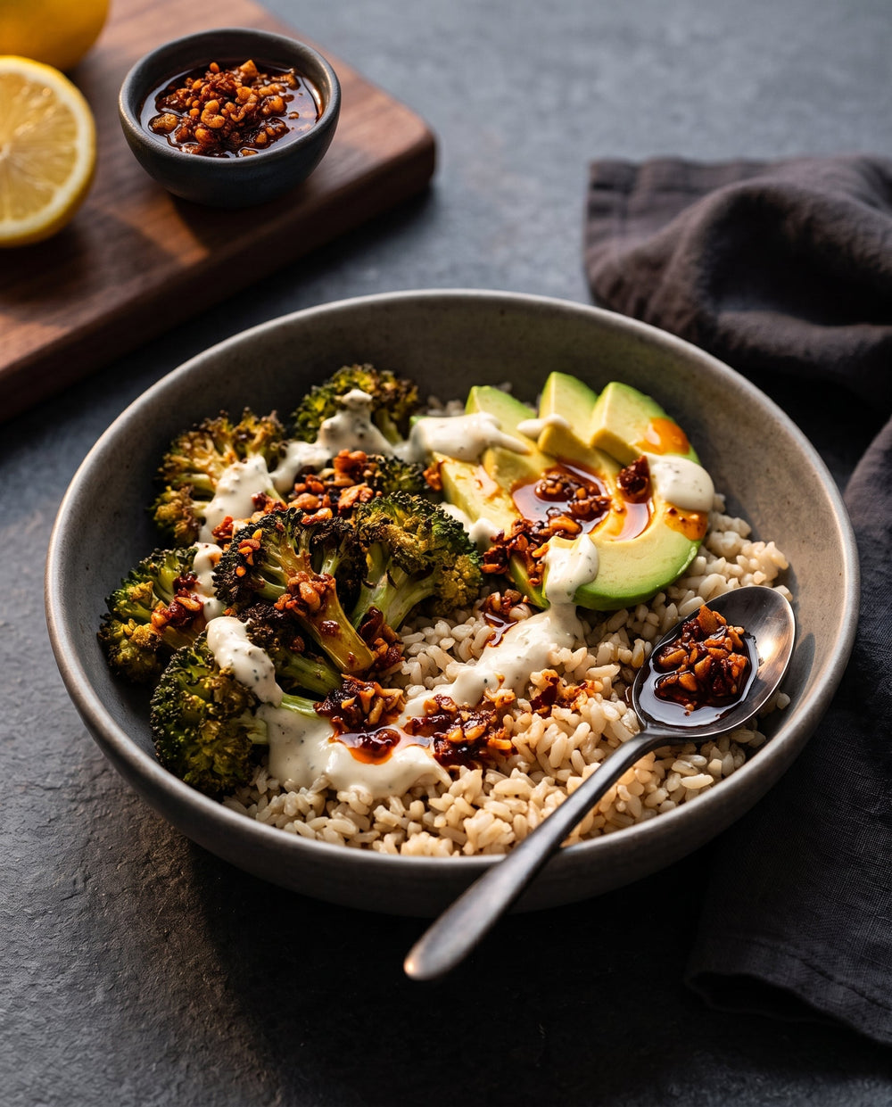 A grey bowl of brown rice topped with charred broccoli, sliced avocado, yogurt sauce, and chili crisp, resting on a dark stone surface with a spoon.