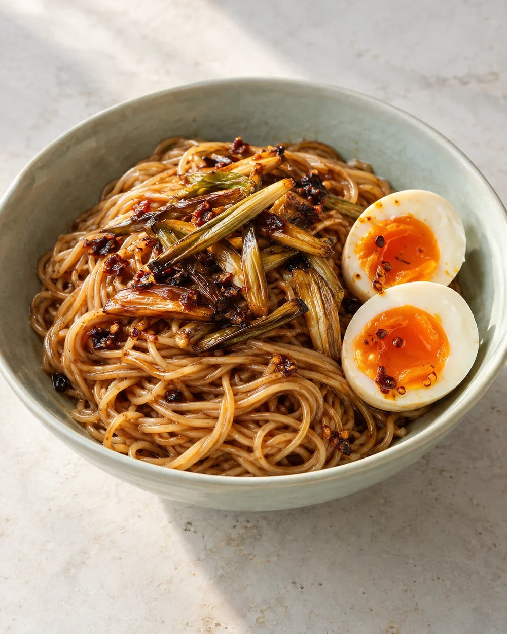 Thin glossy noodles with browned scallions, two jammy egg halves and red-brown chili-crisp solids in a pale green bowl