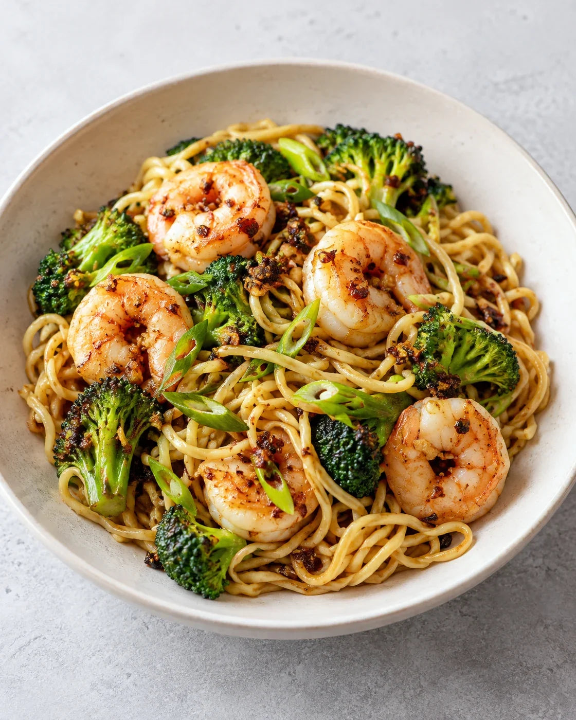 Egg noodles tossed with opaque pink shrimp, browned broccoli, scallions and chili-crisp solids in a shallow ivory bowl
