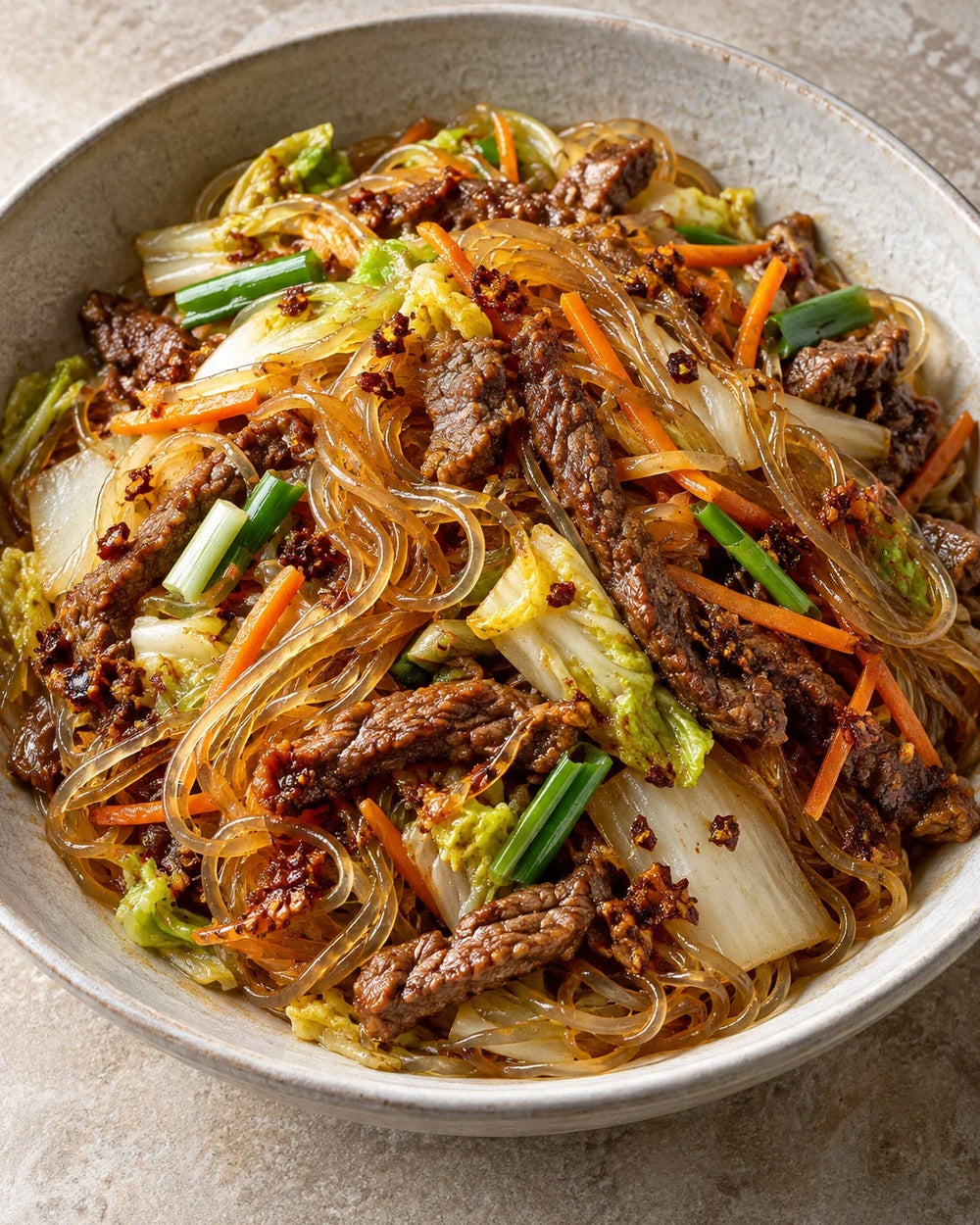 Glossy glass noodles with browned beef strips, napa cabbage, carrot matchsticks, scallions, and spicy chili crisp