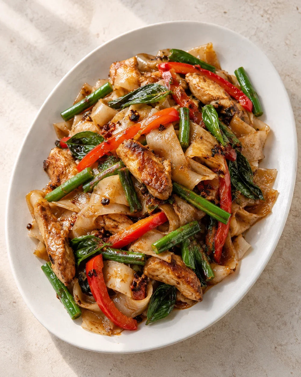 Wide rice noodles with browned chicken, red pepper, green beans, Thai basil and visible chili crisp on a white plate